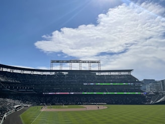 A baseball stadium with a clear sky above. The seating areas are filled with spectators. The field is well-maintained and several players are positioned on it. The scoreboard is visible in the background.
