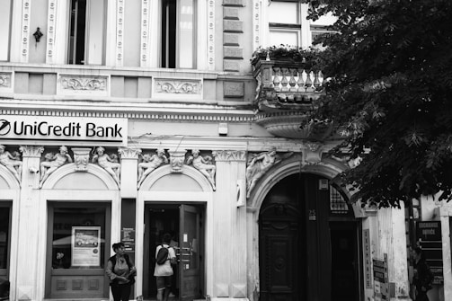 A classic, ornate building facade features intricate architectural details with decorative sculptures and arched entrances. The sign for UniCredit Bank is displayed prominently above the main doorway. Two people stand near the entrance, one carrying a backpack and the other wearing a hat. Lush trees border the building on the right side.