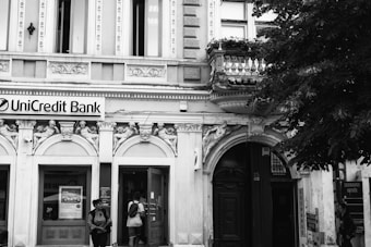 A classic, ornate building facade features intricate architectural details with decorative sculptures and arched entrances. The sign for UniCredit Bank is displayed prominently above the main doorway. Two people stand near the entrance, one carrying a backpack and the other wearing a hat. Lush trees border the building on the right side.