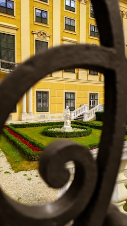 A beautifully manicured garden with neatly trimmed hedges and a circular flower bed featuring bright red and white flowers. A classical statue stands as a focal point. The background shows a yellow building with multiple windows and green shutters, framed by an ornate black wrought iron railing.