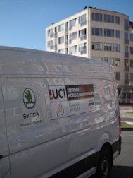 A white van prominently displays logos and information related to the 2021 Road World Championships held in Flanders, Belgium. The van features branding from Skoda as the official car supplier. In the background, there is a modern multi-story building with large windows.