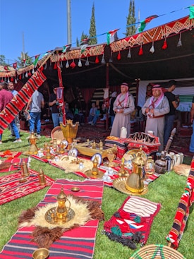 A traditional outdoor setting features colorful textiles, brass coffee pots, and other cultural items arranged on grass. Two men wearing traditional attire stand in front of a tent adorned with intricate patterns. Several visitors are present, engaging with the atmosphere.