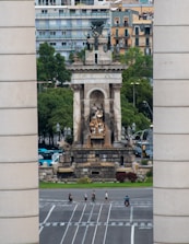 A large monument with intricate sculptures stands prominently in an urban setting. The structure is framed by two tall cylindrical columns, and behind it are several modern apartment buildings. Trees surround the monument, adding greenery to the scene. Below, pedestrians and a person on a motorcycle cross a wide, multi-lane road.