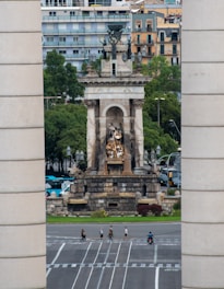 A large monument with intricate sculptures stands prominently in an urban setting. The structure is framed by two tall cylindrical columns, and behind it are several modern apartment buildings. Trees surround the monument, adding greenery to the scene. Below, pedestrians and a person on a motorcycle cross a wide, multi-lane road.
