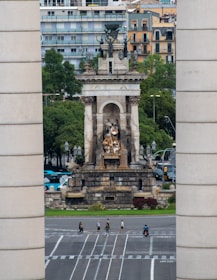 A large monument with intricate sculptures stands prominently in an urban setting. The structure is framed by two tall cylindrical columns, and behind it are several modern apartment buildings. Trees surround the monument, adding greenery to the scene. Below, pedestrians and a person on a motorcycle cross a wide, multi-lane road.