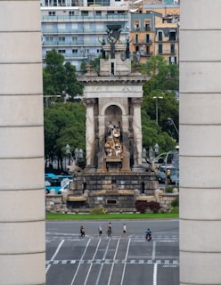 A large monument with intricate sculptures stands prominently in an urban setting. The structure is framed by two tall cylindrical columns, and behind it are several modern apartment buildings. Trees surround the monument, adding greenery to the scene. Below, pedestrians and a person on a motorcycle cross a wide, multi-lane road.