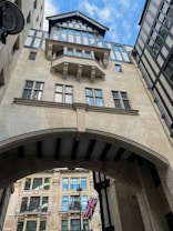 A detailed architectural facade displaying a Tudor-style upper section with exposed beams and multiple windows. Below is a stone archway leading to a space between buildings. A British flag is prominently displayed, hanging from a post, while the sky is a bright blue with some clouds.
