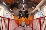 A young child stands in a corridor of a museum or exhibit, gazing at a large satellite model or space-related machinery. The setting is filled with educational displays and blue informational panels on the walls. A red railing lines the path, adding a vibrant contrast to the metallic and orange hues of the machinery.
