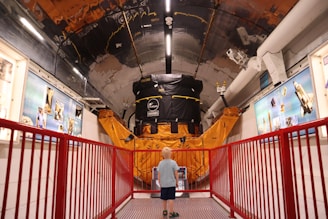 A young child stands in a corridor of a museum or exhibit, gazing at a large satellite model or space-related machinery. The setting is filled with educational displays and blue informational panels on the walls. A red railing lines the path, adding a vibrant contrast to the metallic and orange hues of the machinery.