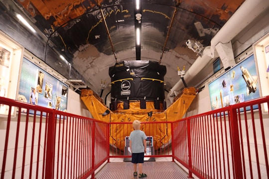 A young child stands in a corridor of a museum or exhibit, gazing at a large satellite model or space-related machinery. The setting is filled with educational displays and blue informational panels on the walls. A red railing lines the path, adding a vibrant contrast to the metallic and orange hues of the machinery.