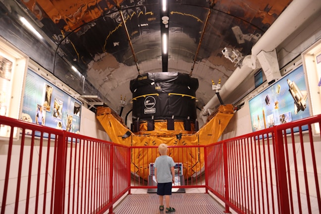 A young child stands in a corridor of a museum or exhibit, gazing at a large satellite model or space-related machinery. The setting is filled with educational displays and blue informational panels on the walls. A red railing lines the path, adding a vibrant contrast to the metallic and orange hues of the machinery.