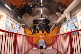 A young child stands in a corridor of a museum or exhibit, gazing at a large satellite model or space-related machinery. The setting is filled with educational displays and blue informational panels on the walls. A red railing lines the path, adding a vibrant contrast to the metallic and orange hues of the machinery.