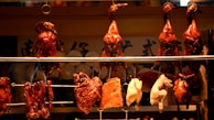 A row of roasted ducks hangs in a display window, accompanied by other types of cooked meat. The ducks are a glossy brown color, indicating they are heavily glazed or roasted. Some other meat cuts are also on display below the ducks.