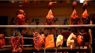 A row of roasted ducks hangs in a display window, accompanied by other types of cooked meat. The ducks are a glossy brown color, indicating they are heavily glazed or roasted. Some other meat cuts are also on display below the ducks.