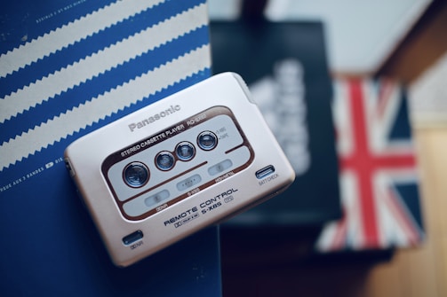 A vintage Panasonic stereo cassette player with metallic buttons lies on a striped blue background, next to a partially visible British flag. The device features buttons for play, stop, and other functions typical of a cassette player.