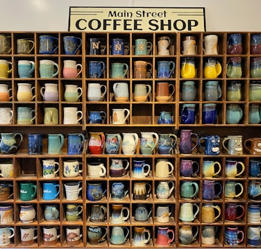A wooden shelf filled with an assortment of colorful, handcrafted mugs. Each cubby hole contains a unique mug with various patterns and colors, including blues, greens, yellows, and reds. Above the shelves, a sign reads 'Main Street Coffee Shop.' The overall display is artistic and visually appealing, showcasing a variety of designs and textures.