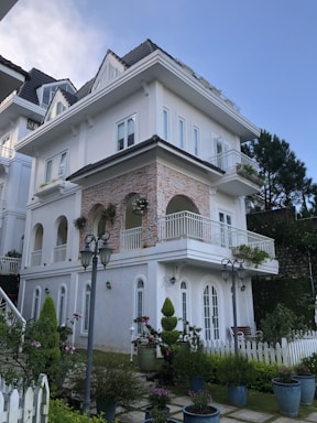 A large, elegant white house with three stories and a dark grey tiled roof. The facade features arched windows and balconies adorned with potted plants and columns, creating a sophisticated appearance. The surrounding area includes a well-maintained garden with various shrubs and flowers, and a white picket fence encircles the property.