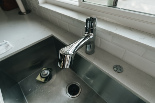 A modern kitchen sink featuring a sleek stainless steel faucet, a deep metallic basin with a drain, and a reflective surface. The countertop is made of white marble, and a window above the sink allows natural light to illuminate the area. A sponge rests at the bottom of the sink, indicating recent use.