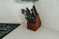 A neatly organized set of kitchen knives is placed in a wooden knife block on a marble countertop. The background features white subway tiles and there is a stovetop with black grates to the left. There are also some wooden utensils in a container near the knife block.