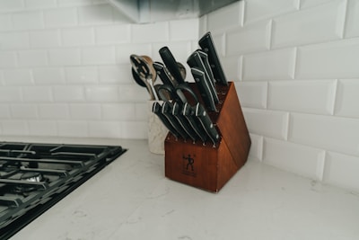 A neatly organized set of kitchen knives is placed in a wooden knife block on a marble countertop. The background features white subway tiles and there is a stovetop with black grates to the left. There are also some wooden utensils in a container near the knife block.