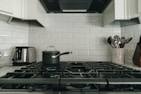 A modern kitchen with a gas stove featuring five burners, a pot with a lid on one of the burners, a stainless steel toaster on the left, and a utensil holder with various kitchen utensils on the right. The background consists of white subway tiles.