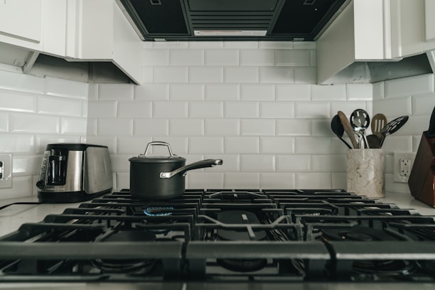 A modern kitchen with a gas stove featuring five burners, a pot with a lid on one of the burners, a stainless steel toaster on the left, and a utensil holder with various kitchen utensils on the right. The background consists of white subway tiles.