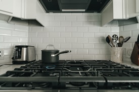 A modern kitchen with a gas stove featuring five burners, a pot with a lid on one of the burners, a stainless steel toaster on the left, and a utensil holder with various kitchen utensils on the right. The background consists of white subway tiles.