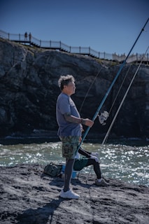 A man stands on a rocky shoreline, holding a fishing rod poised above the water. He is dressed in casual outdoor attire with camouflage shorts and a blue shirt. Behind him, a cliff and a fence are visible with a couple of people in the distance observing the scene. The sky is clear and the water beneath reflects the sunlight.