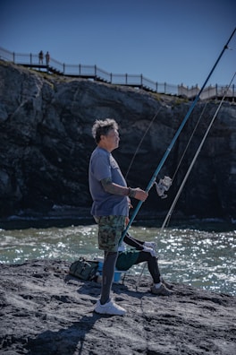A man stands on a rocky shoreline, holding a fishing rod poised above the water. He is dressed in casual outdoor attire with camouflage shorts and a blue shirt. Behind him, a cliff and a fence are visible with a couple of people in the distance observing the scene. The sky is clear and the water beneath reflects the sunlight.