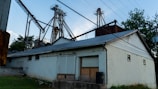 An industrial facility with a large metal structure featuring tall towers and connecting pipes. The building in the foreground is constructed from white-painted bricks and has a metal roof. Surrounding the area are trees and grass, while power lines stretch across the sky.