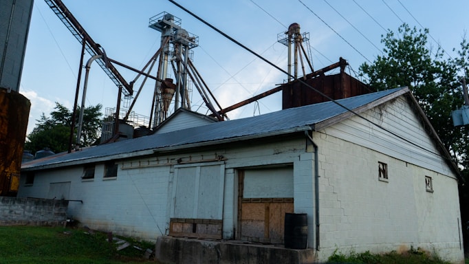 An industrial facility with a large metal structure featuring tall towers and connecting pipes. The building in the foreground is constructed from white-painted bricks and has a metal roof. Surrounding the area are trees and grass, while power lines stretch across the sky.