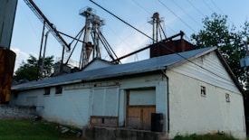 An industrial facility with a large metal structure featuring tall towers and connecting pipes. The building in the foreground is constructed from white-painted bricks and has a metal roof. Surrounding the area are trees and grass, while power lines stretch across the sky.
