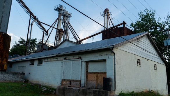 An industrial facility with a large metal structure featuring tall towers and connecting pipes. The building in the foreground is constructed from white-painted bricks and has a metal roof. Surrounding the area are trees and grass, while power lines stretch across the sky.