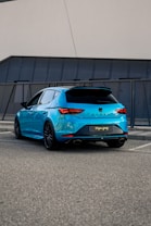 A bright blue hatchback car is parked in a modern urban setting, with sleek, angular design lines and black-tinted windows. The car features prominent dual exhaust pipes and distinctive rear light shapes. It is situated on a gray asphalt surface near a contemporary building facade with textured panels.