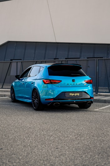 A bright blue hatchback car is parked in a modern urban setting, with sleek, angular design lines and black-tinted windows. The car features prominent dual exhaust pipes and distinctive rear light shapes. It is situated on a gray asphalt surface near a contemporary building facade with textured panels.