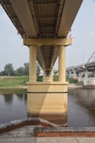 A large, concrete bridge extends over a body of water with multiple supporting pillars visible. The bridge is situated in an urban environment, with a river running below and greenery along the banks. Nearby infrastructure and surrounding landscape indicate an area likely within a city.