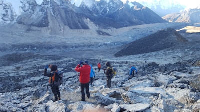 Several hikers are trekking along a rocky, mountainous path surrounded by snow-capped peaks. The landscape is rugged and expansive, with layers of distant mountains. The group wears colorful hiking gear, including jackets and backpacks.