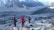 Several hikers are trekking along a rocky, mountainous path surrounded by snow-capped peaks. The landscape is rugged and expansive, with layers of distant mountains. The group wears colorful hiking gear, including jackets and backpacks.