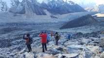 Several hikers are trekking along a rocky, mountainous path surrounded by snow-capped peaks. The landscape is rugged and expansive, with layers of distant mountains. The group wears colorful hiking gear, including jackets and backpacks.