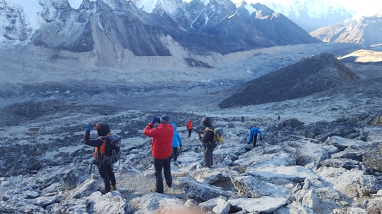 Several hikers are trekking along a rocky, mountainous path surrounded by snow-capped peaks. The landscape is rugged and expansive, with layers of distant mountains. The group wears colorful hiking gear, including jackets and backpacks.