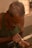 An elderly man is intently working on a small wooden object using his hands. His focus is on the fine details of the craftsmanship, displaying his skill and patience. The background is softly blurred, highlighting the concentration on his task.