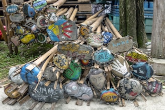 A pile of bamboo sticks with attached oval-shaped wooden signs, each featuring unique and colorful hand-painted designs and text in various languages. The signs display a variety of artistic styles, including abstract patterns, text, handprints, and faces. They rest against a tree and are stacked compactly, with greenery visible in the background.