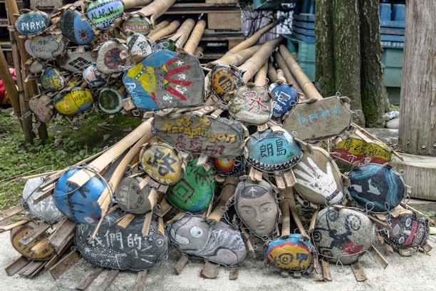 A pile of bamboo sticks with attached oval-shaped wooden signs, each featuring unique and colorful hand-painted designs and text in various languages. The signs display a variety of artistic styles, including abstract patterns, text, handprints, and faces. They rest against a tree and are stacked compactly, with greenery visible in the background.