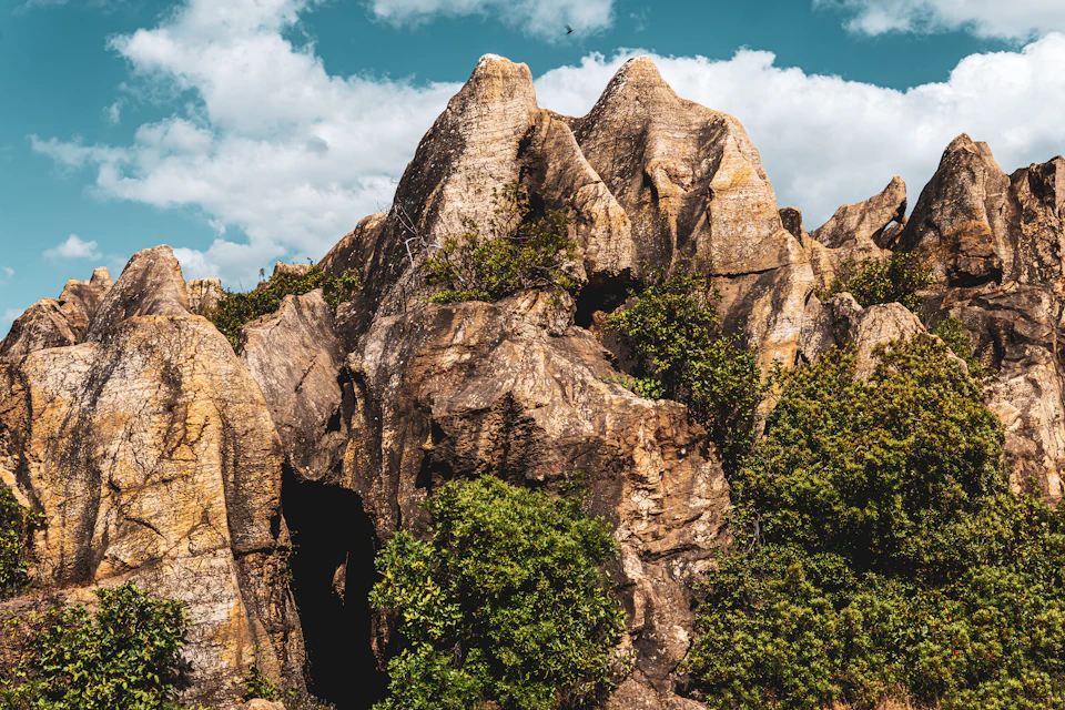 The image depicts a rocky cliff face with numerous large, jagged rocks and boulders in varying shades of brown and gray. The cliff is covered in lush green vegetation, including trees and bushes that add color to the otherwise monochromatic landscape. A small cave can be seen on one side of the cliff, providing a glimpse into the rugged terrain below. The sky above is clear blue with scattered clouds, creating a contrast between the rocky cliffs and the expansive sky.