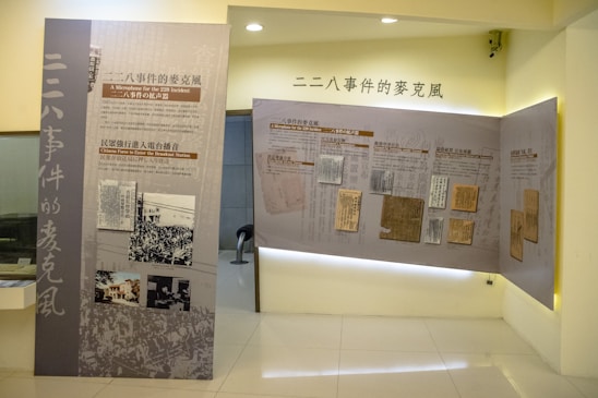 A museum exhibit features large panels with images and text related to the 228 Incident, displayed in a well-lit, modern room. One panel includes news clippings and historical photographs, while the other displays documents. The exhibit is presented in both Chinese and English.