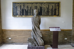 A statue of a man in a suit stands against a brick wall. Above the statue, there is a large black plaque with gold-colored inscriptions in Chinese characters. To the right of the statue is a pedestal with an information board containing text and images.