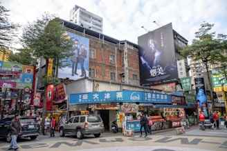 The image depicts a busy urban street scene featuring various shops, billboards, and advertisements. Two large billboards hang on the side of an old, multi-story brick building. One of the billboards has an image of two people in a dramatic pose, while the other shows a person with an intense look. There are several people walking on the street or standing near the shops. Cars are parked nearby and are moving along the road. The street is adorned with colorful signage, posters, and has a mixture of modern and older architectural elements.