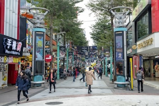 A busy urban street scene with people walking, storefronts, and advertising signs in an area that appears to be a shopping district. The street is lined with colorful signage and various shops, including international brands. Tall trees are present along the sides, and the area has an energetic atmosphere with people dressed in winter clothing.