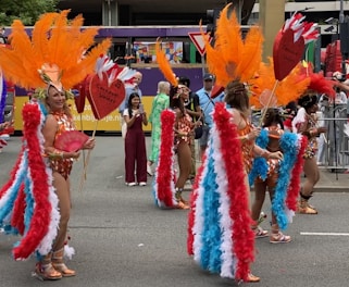 A group of performers in colorful costumes parades through a street. The costumes feature large, vibrant feather headdresses in orange, red, and white, along with matching feathered wings. They carry signs shaped like red hearts. Spectators are visible in the background observing the parade.