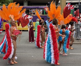A group of performers in colorful costumes parades through a street. The costumes feature large, vibrant feather headdresses in orange, red, and white, along with matching feathered wings. They carry signs shaped like red hearts. Spectators are visible in the background observing the parade.